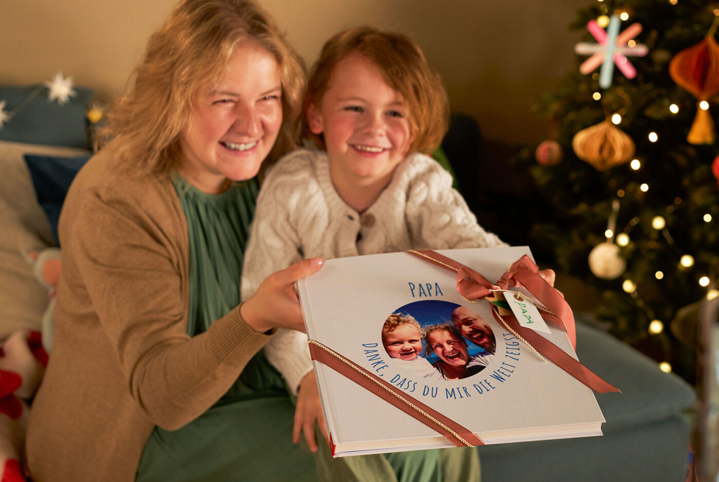 Eine Frau sitzt mit ihrem Kind vor einem Weihnachtsbaum. Beide lachen und halten ein CEWE FOTOBUCH in den Händen.